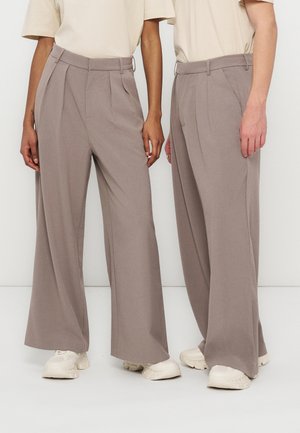 Two people wearing beige wide-leg trousers, cream sneakers, and light beige short-sleeve tops, standing side by side against a white background.