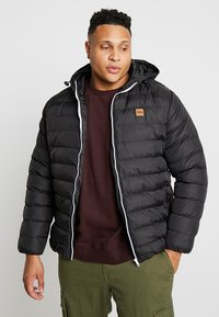 Sort pufferjakke med horisontal quiltning, hvid lynlås og en brun læderlap. Jakken har en hætte og en afslappet pasform.