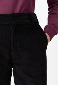 Pantalones de pana negros con corte recto, que presentan estrías pronunciadas, bolsillos laterales y una cinturilla sólida. Combinados con una blusa de manga larga color burdeos.