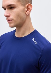 Άνδρας που φορά μπλε μπλουζάκι με στρογγυλή λαιμόκοψη και λευκό κείμενο "BORG" κοντά στον δεξί ώμο, κοιτάζει αριστερά με ουδέτερη έκφραση, κοντά μαλλιά και σκουλαρίκια.