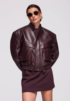 Vrouw draagt een oversized donkerpaarse lederen jas, bijpassende rok, grote gouden oorbellen en een zonnebril, poseert met handen achter de rug.