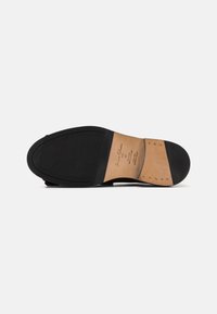 Suela de zapato de cuero negro con superficie de goma estriada, diseño curvado y acentos de cuero beige que presentan costuras visibles y detalles de marca.
