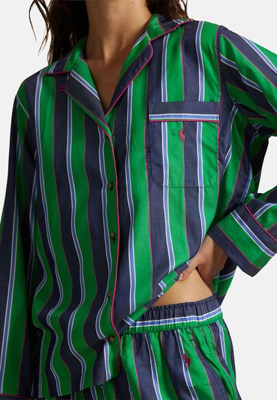 Frau trägt einen grün-, marine- und blau gestreiften Pyjamaoberteil und eine Schlafanzughose mit rosa Paspelierung und besticktem Logo auf der Tasche und den Shorts.