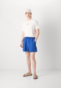 Camiseta blanca con el logo "GANT", pantalones cortos azules con textura y cinturilla elástica, y sandalias beige con dos tiras, de pie sobre un fondo blanco.