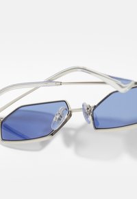 Ochelari de soare hexagonali cu lentile albastre, ramă metalică argintie, pernuțe pentru nas transparente și brațe curbe, evidențiind un design minimalistic.