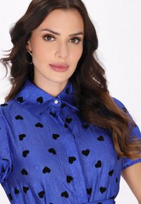 Camicia blu con colletto, caratterizzata da una texture stropicciata e da motivi a cuori neri sparsi. Include un dettaglio decorativo con bottoni.
