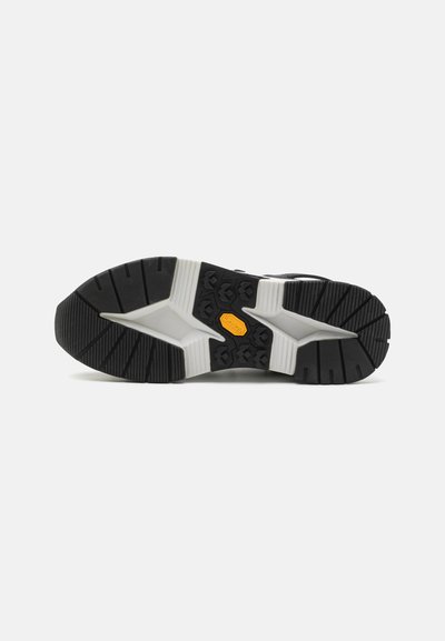 Musta kumipohja, jossa on valkoinen välipohja. Ominaisuuksiin kuuluvat syvät urat ja keltainen Vibram-logo, jotka on suunniteltu vakautta ja pitoa varten.