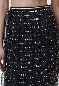 Jupe en tulle noire avec des pois blancs, un design plissé et une taille accentuée en or. La texture est transparente et légère.