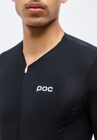 Κοντινό πλάνο άνδρα που φοράει μαύρο ποδηλατικό τζέρσεϊ με φερμουάρ και λευκό λογότυπο "poc" στο αριστερό μέρος του στήθους.