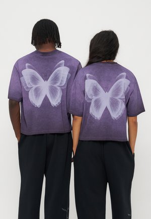 Due persone indossano camicie viola con grandi stampe bianche di farfalle sulla schiena, abbinate a pantaloni neri, viste da dietro.