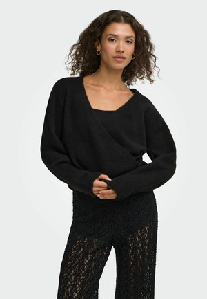 Femme aux cheveux bouclés portant un pull noir à col en V, un top noir en dessous et un pantalon en dentelle noire texturée, debout devant un fond uni.