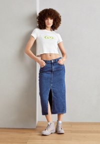 Kvinde med krøllet hår iført en hvid crop top med et grønt logo, blå midi denimnederdel med forslids og beige støvler, som står indendørs.