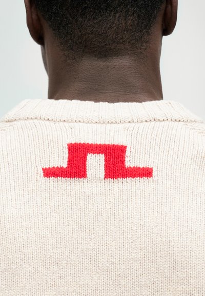 Bežinė megzta palaidinė su raudonu blokiniu raštu nugaroje. Audinys yra tekstūruotas, matomi siuvimo siūlai ir vangus apykaklė.