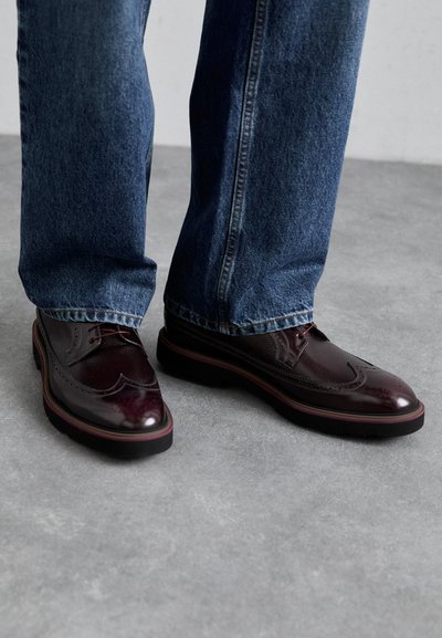 Brogues in pelle borgogna con perforazioni decorative, abbinati a jeans in denim blu scuro. Suole spesse nere con un sottile bordo colorato.
