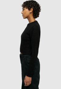 Maglione corto nero con un design a maglia testurizzata, scollatura tonda e maniche lunghe, abbinato a pantaloni scuri. Vista di profilo.