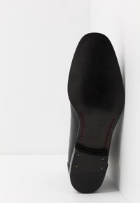 Semelle de chaussure en cuir noir avec une finition lisse, bout pointu, détails de couture rouges et clous d'accent dorés près du talon. Conçue pour une tenue formelle.
