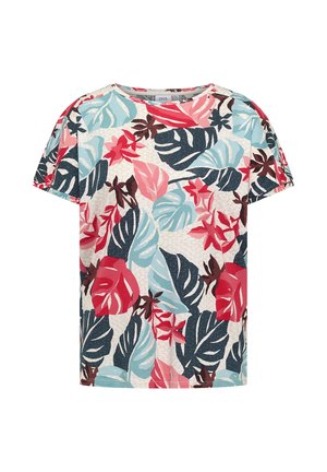 Femme debout souriante portant un haut à manches courtes fleuri multicolore, un jean à jambes larges bleu clair et des baskets beiges.