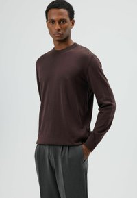 Pull marron à manches longues en matière douce avec un col rond, associé à un pantalon gris sur mesure. Le design présente une coupe décontractée.