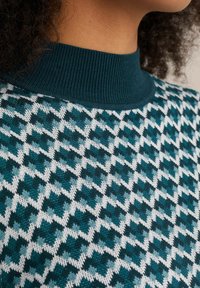 Sweter dziergany w turkusowo-białym geometrycznym wzorze, z ribbowanym turkusowym kołnierzem i miękką teksturą. Zbliżenie ukazujące szczegóły wzoru.