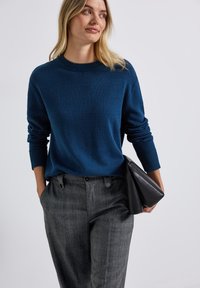 Pull en maille bleu marine à col rond et poignets côtelés, associé à un pantalon à carreaux gris et un sac à main noir.