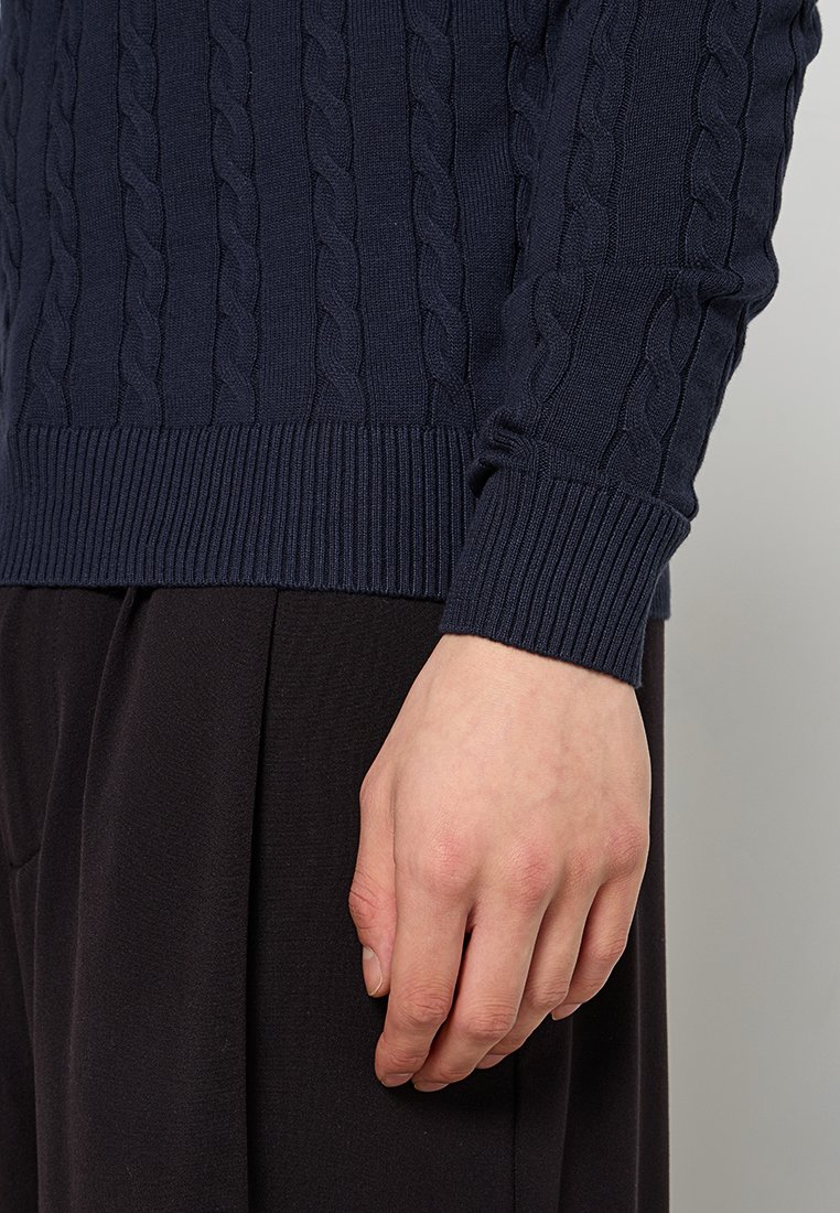 Hand, die neben dem Körper ruht, trägt einen navyblauen, strukturierten Pullover und schwarze Hosen, sichtbar vom Taillenbereich bis zu den Fingern.