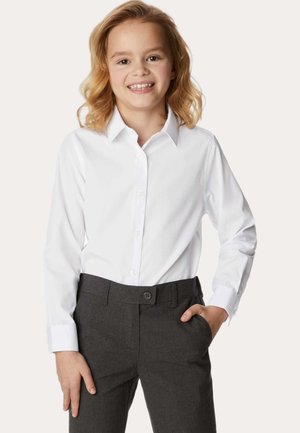 Lächelndes Kind mit schulterlangem blonden Haar, das ein weißes, knöpfbares Hemd und dunkelgraue Hosen trägt, mit einer Hand in der Tasche.