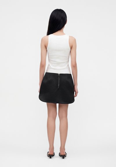 Femme debout, dos tourné, portant un débardeur blanc, une mini-jupe noire avec une fermeture éclair dans le dos et des sandales à talons noires sur fond gris.
