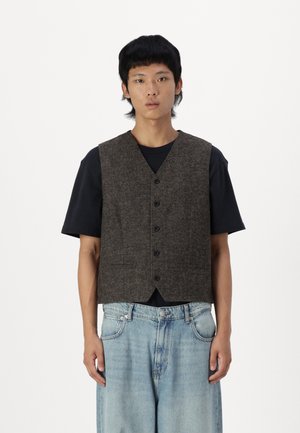 Gilet en laine marron avec une finition texturée, cinq boutons et un design col en V, porté sur un t-shirt noir et assorti à un pantalon en denim clair.