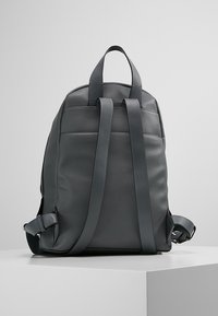 Sac à dos en cuir gris avec des bretelles réglables, une poignée supérieure, une texture lisse et un design épuré. Présente un détail de couture à l'arrière.