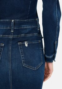 Zoom su una persona che indossa una giacca di jeans blu scuro e una gonna, con tasca posteriore visibile e dettagli in metallo argentato sulla tasca.