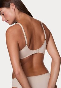 Soutien-gorge beige à armatures avec bretelles réglables, présentant une texture lisse, une fermeture à crochets et œillets, et des accents en métal doré.