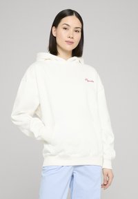 Sudadera con capucha crema con un bolsillo canguro delantero, puños y dobladillo acanalados, con texto bordado en rojo en el pecho. Combinada con pantalones a rayas azul claro.