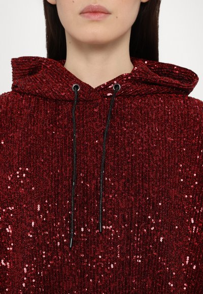 Red sequin hoodie with a textured finish, featuring a black drawstring and hood. Positioned at the neckline, the sequins create a shimmering effect.