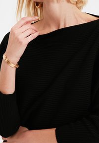 Zwarte geribbelde trui met een off-shoulder ontwerp. Heeft een gladde stoftextuur. Een gouden armband en oorbellen voegen opmerkelijke metallic accenten toe.