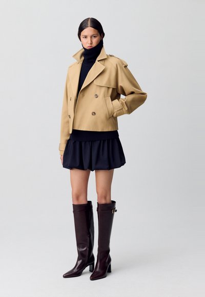 Veste cropped beige à double boutonnage avec grands revers, associée à un col roulé noir et une jupe volantée bleu marine. Bottes bordeaux montantes.
