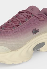 Κοντινό πλάνο ενός ροζ και λευκού sneakers με διαβάθμιση, υφασμάτινο επάνω μέρος με υφή, παχύ παχύρευστο πέλμα κρεμ, κορδόνια και ένα μικρό κεντημένο λογότυπο κροκόδειλου στο πλάι.