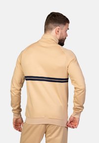 Beige lynlåsjakke med høj krave, med en marineblå streg på tværs af ryggen. Glat tekstur og tætsiddende design. Ribstrikkede manchetter og kant.