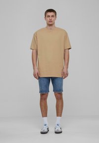 T-shirt en coton beige oversize à manches courtes, associé à un short en denim bleu. Le modèle porte des chaussettes blanches et des baskets grises avec des rayures noires.