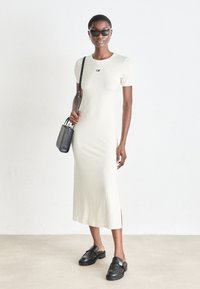 Light yellow form-fitting dress with short sleeves, side split, embroidered logo. Paired with black bag and flat black shoes.