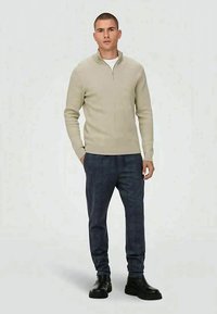 Young man standing, wearing a beige quarter-zip sweater, dark plaid pants, and black leather boots, hands relaxed by sides.