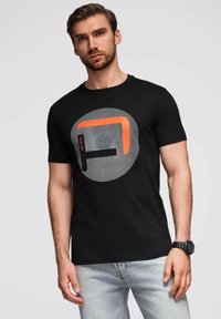 Černé bavlněné tričko s kulatým grafickým designem v oranžových a šedých akcentách. Model má na sobě světle šedé džíny a černé hodinky.