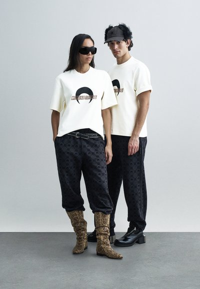 White t-shirts with black crescent logo, black patterned pants, brown snakeskin boots, and black shoes. Accessories include dark sunglasses and a fuzzy cap.