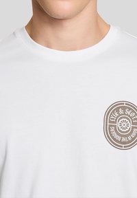 Άνδρας που φοράει λευκό T-shirt με στρογγυλή λαιμόκοψη και μικρό στρογγυλό καφέ λογότυπο στο αριστερό στήθος με την επιγραφή "Lyle & Scott Born in the Borders".