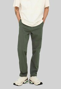 Persona con pantalones verde oliva, zapatillas blancas y una camiseta de manga corta color crema, con las manos en los bolsillos frente a un fondo sencillo.