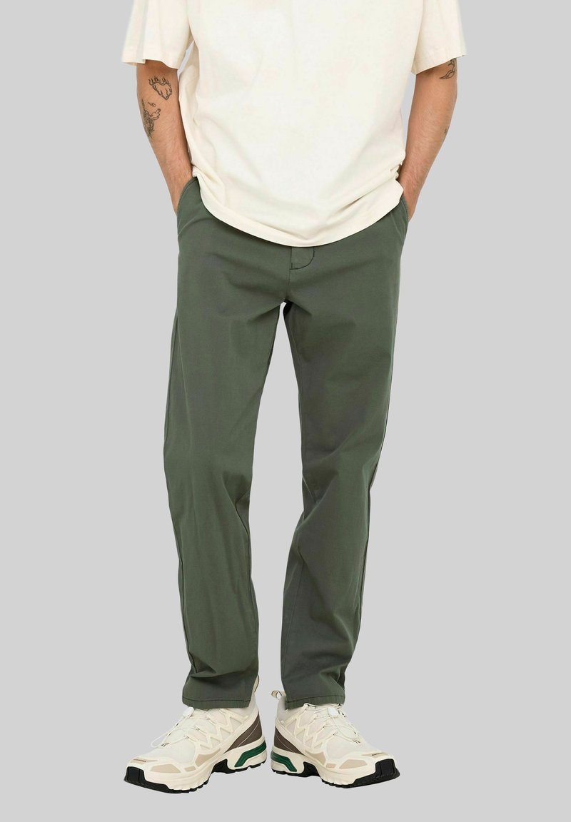 Persona con pantalones verde oliva, zapatillas blancas y una camiseta de manga corta color crema, con las manos en los bolsillos frente a un fondo sencillo.