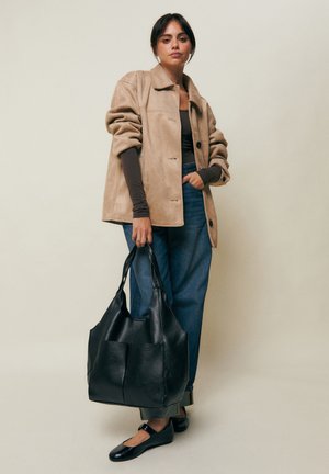 Naine, kes kannab beeži jopet, tumedat pika varrukaga pluusi, siniseid teksapükse ja musti madalaid jalanõusid, hoides suurt musta nahast käekotti, seisab ühevärvilise tausta ees.