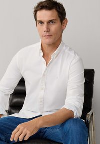 Man met kort bruin haar, gekleed in een wit overhemd met knopen en een blauwe spijkerbroek, zittend op een zwarte leren stoel tegen een effen achtergrond.