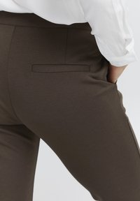 Hnedé slim-fit nohavice z hladkej tkaniny, s vrchnou zadnou vreckom a jemným prešívaním, v kombinácii s bielou košeľou.