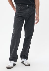 Hombre que lleva jeans rectos de mezclilla deslavados negros y zapatillas blancas con rayas negras, de pie con una mano en el bolsillo.