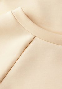 Béžová tkanina s hladkou, mírně lesklou texturou. Má kulatý výstřih a švové detaily, které ukazují minimalistický design.